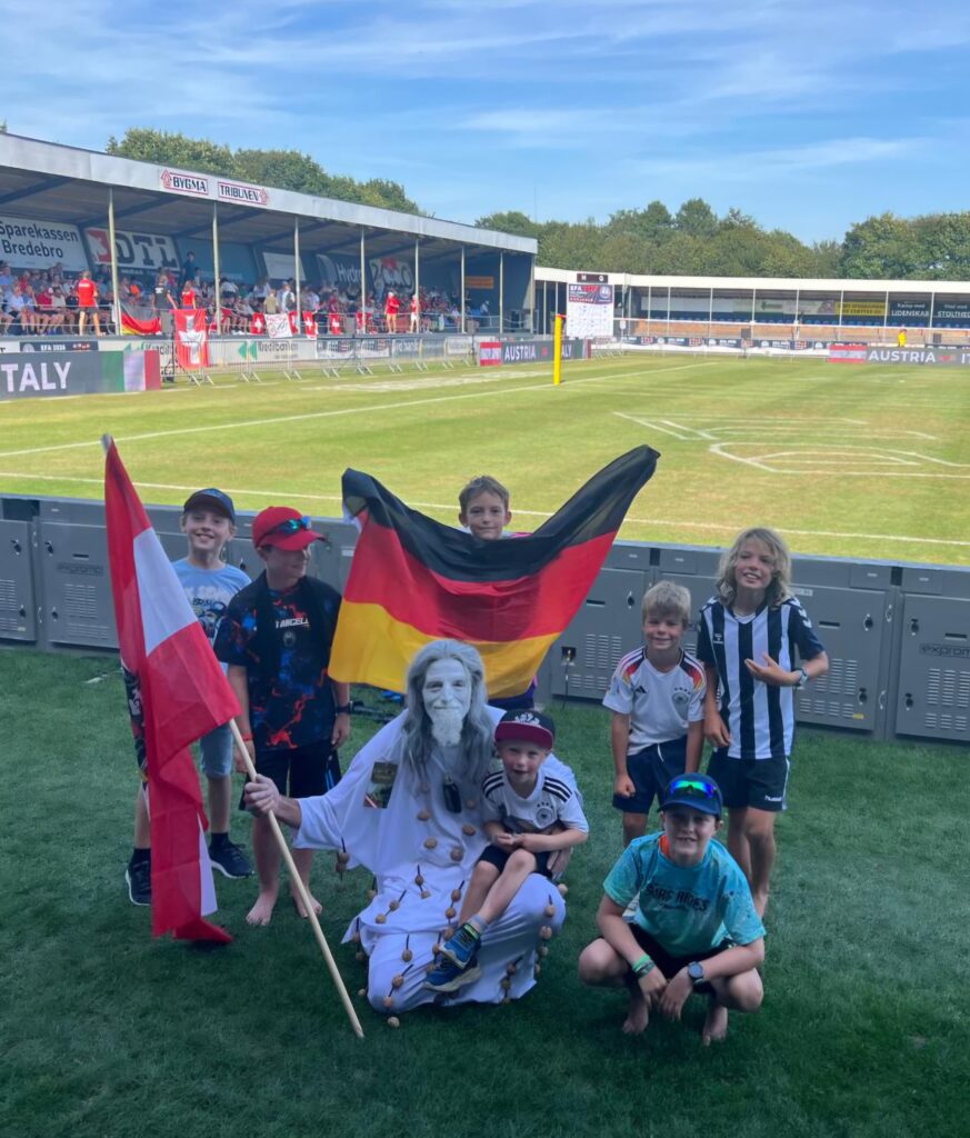 Schülper Kinder mit Deutschland- und Österreichfahnen sowie dem Nussgeist am Spielfeldrand in Tondern