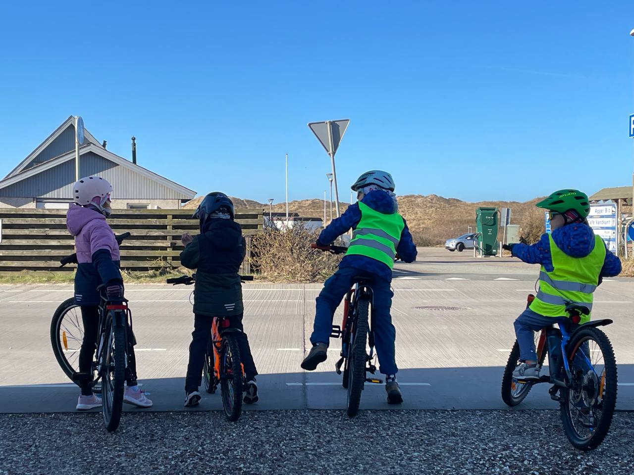 Vier Kinder stehen mit Fahrrädern und Helmen an einer Kreuzung, zwei von ihnen tragen gut sichtbare Warnwesten