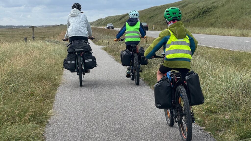 Ein Erwachsener und zwei Kinder fahren mit Fahrradhelmen, Warnwesten und Gepäcktaschen auf einem Radweg in Dänemark